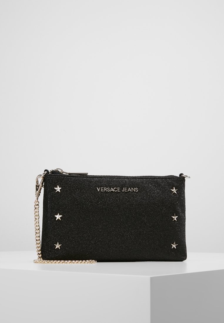 Pochette noire pailletée avec des accents d'étoiles argentées, dotée d'une fermeture éclair et d'une bandoulière chaîne amovible. Nom de la marque "VERSACE JEANS" visible.