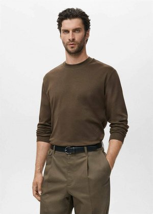 Bruine langemouwen shirt in een zachte stof, ronde halslijn, gecombineerd met lichtbruine broek en een zwarte riem, model staat met handen in de zakken.