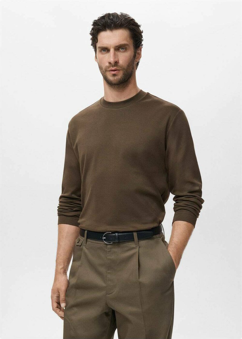 Smeđa košulja dugih rukava od mekog materijala, okrugli izrez, u kombinaciji sa svijetlosmeškim hlačama i crnim remenom, model stoji s rukama u džepovima.