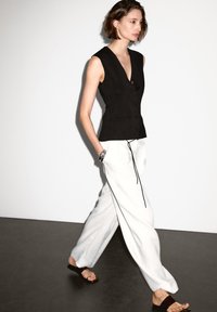 Zwarte mouwloze vest met knopen, gecombineerd met losse witte broeken met een zwarte trekkoord. Model draagt zwarte sandalen en een zilveren armband.