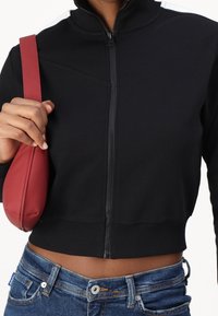 Schwarze, kurz geschnittene Jacke mit Reißverschluss und hohem Kragen, mit strukturierter Oberfläche und weißen Akzenten an den Schultern, kombiniert mit einer kleinen roten Handtasche.