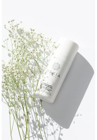 Bottiglia bianca cilindrica etichettata "OSKIA Renaissance Cleansing Gel" con un logo rotondo, posizionata tra delicati fiori bianchi su una superficie luminosa.