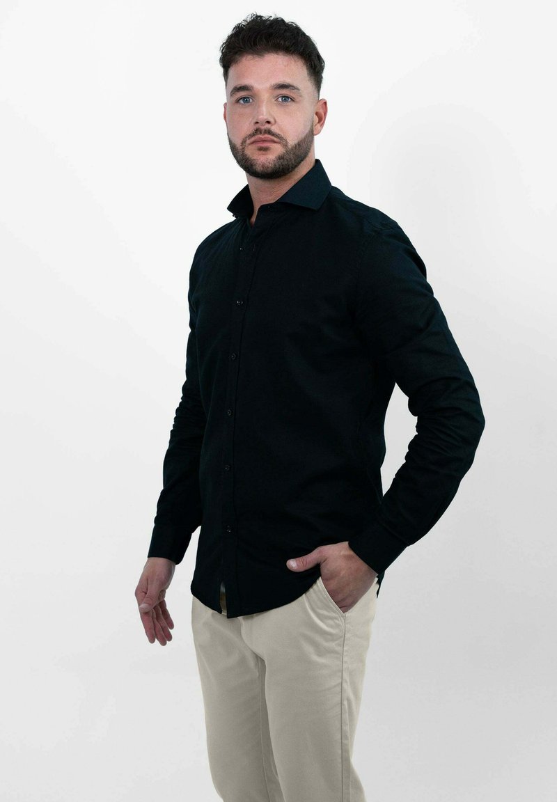 Zwarte langemouwen shirt met knopen, kraag en aansluitend ontwerp, gecombineerd met lichtbeige broek, tegen een effen witte achtergrond.