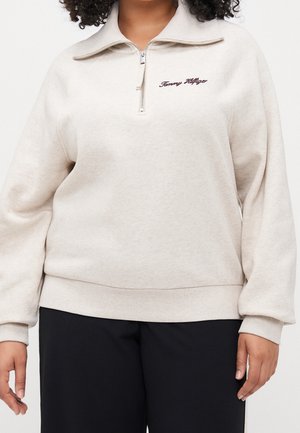 Kobieta ubrana w kremową bluzę z półzamkiem i haftem "Tommy Hilfiger" na piersi oraz czarne spodnie.