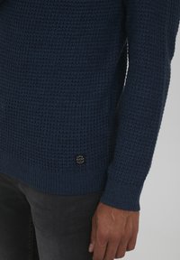 Suéter de punto en azul oscuro con un tejido texturizado, puños y dobladillo acanalados, que presenta un pequeño parche de logotipo negro en la parte inferior izquierda.