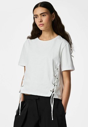 Jeune femme aux longs cheveux noirs portant un haut blanc à manches courtes avec des détails de laçage sur les côtés et un pantalon noir taille haute.