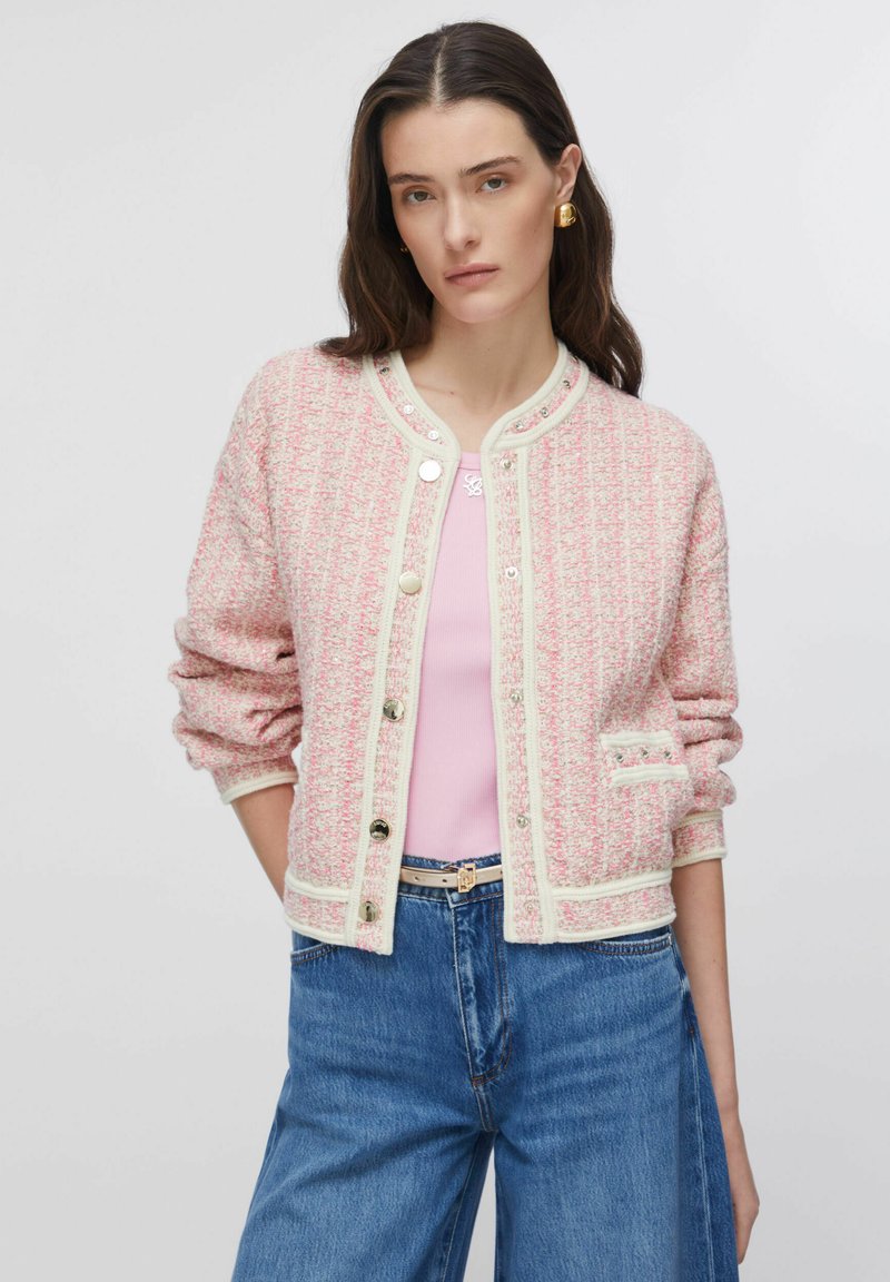 Cárdigan de tweed tejido en rosa y blanco con ribete crema, escote redondo, cierre de botones y bolsillo delantero. Combinado con una camiseta rosa claro y vaqueros azules.