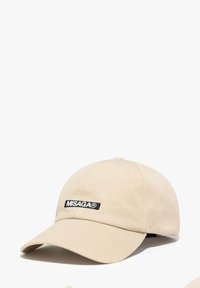 Beżowa czapka bejsbolowa z teksturowanej tkaniny, z wygiębrim daszkiem i czarną naszywką z logo "MISAGA" z przodu, wyposażona w regulowany pasek z tyłu.