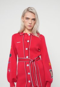 Czerwona dzianinowa sukienka z kołnierzykiem, zapinana na guziki z długimi rękawami. Posiada haftowane akcenty oraz związany pasek w talii.