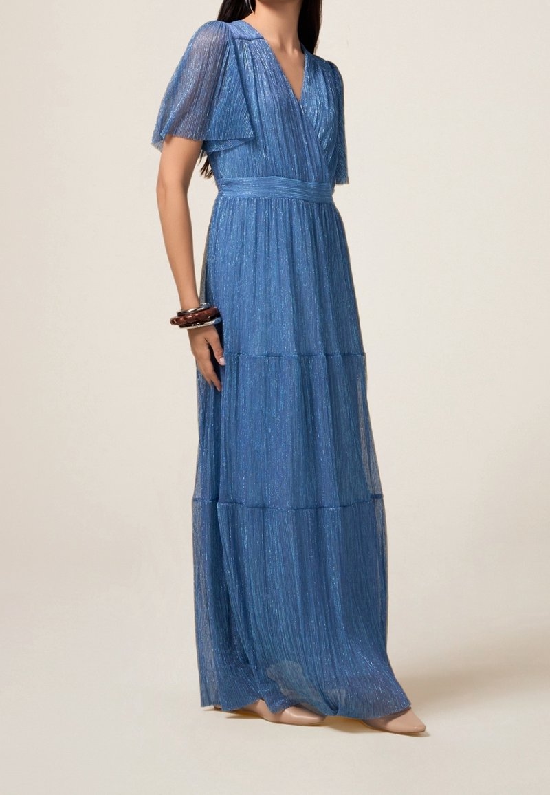 Femme portant une robe bleue scintillante longue jusqu'au sol avec des manches flottantes et un corsage style portefeuille, associée à des chaussures pointues couleur nude et des bracelets.