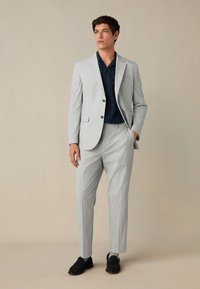 Costume rayé gris clair avec une veste cintrée et un pantalon fuselé, associé à une chemise manches courtes bleu marine. Chaussures : mocassins foncés.