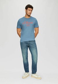 Blaues Baumwoll-T-Shirt mit rotem "s.Oliver"-Logo, kombiniert mit hellblauen Jeans und weißen Sneakern. Das Shirt hat einen Rundhalsausschnitt und kurze Ärmel.
