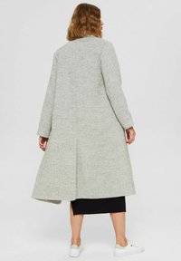 Person with shoulder-length brown hair wearing a long gray textured coat, black skirt, and white sneakers, standing on a plain background.