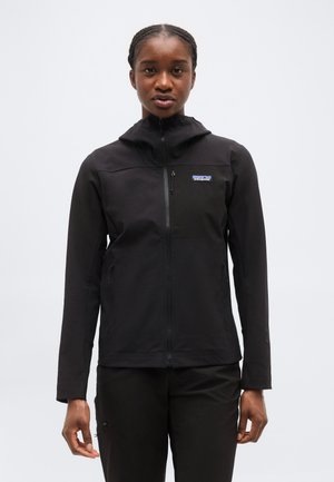 Žena v čiernej bunde na zips s kapucňou a logom Patagonia, stojaca pred jednoduchým svetlým pozadím.