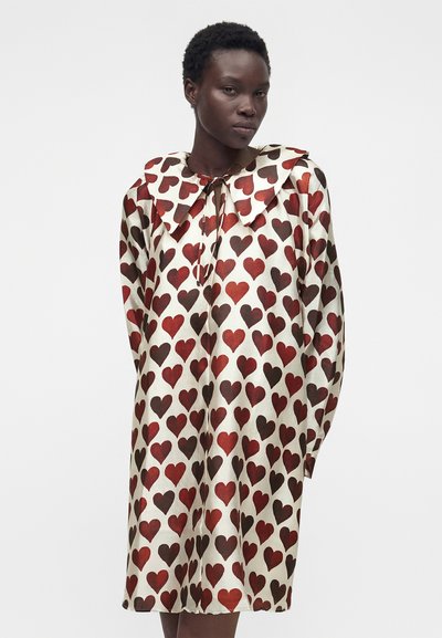 Γυναίκα που φοράει μακρυμάνικο φόρεμα κρεμ χρώματος με μεγάλο γιακά, καλυμμένο με κόκκινα και σκούρα καφέ σχέδια καρδιών, που στέκεται μπροστά σε απλό φόντο.