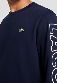 Felpa blu navy in misto cotone, con un piccolo logo coccodrillo ricamato sul petto e scritte bianche in grassetto lungo la manica.