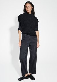 Maglione lavorato a maglia nera con colletto alto, abbinato a jeans grigio scuro a vita alta. Le calzature sono mocassini neri.