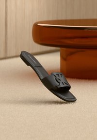 Sandale slide noire Calvin Klein avec design de logo découpé sur un tapis beige, près d'un pied de table rond translucide marron.