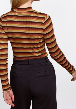 Maglietta a maniche lunghe con texture a coste caratterizzata da strisce orizzontali nei colori marrone, giallo e arancione. Indossata con pantaloni neri su misura.
