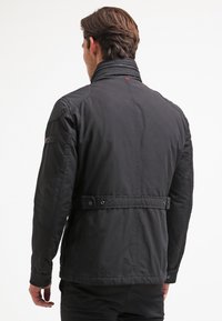 Svart lättviktsjacka med ståkrage, tryckknappsstängning och bälte i midjan. Har texturerad tyg och subtila sidodetaljer.