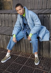 Cappotto imbottito azzurro chiaro sopra una tuta in denim, accessoriodato con una maglietta verde a maniche lunghe e scarpe slip-on a fantasia con suola bianca.