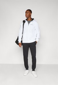 Witte zip-up hoodie met trekkoorden, zwart logo; grijze T-shirt eronder, donkergrijze sweatpants en witte sneakers. Zwarte tas over de schouder.
