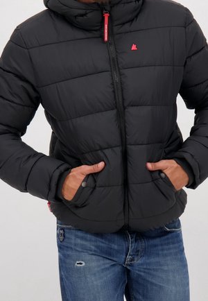 Personne portant une veste matelassée noire avec fermeture éclair frontale et logo rouge, mains dans les poches, associée à un jean bleu déchiré.