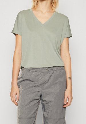 Personne portant un haut vert clair à manches courtes et col en V ainsi qu'un pantalon gris froissé, avec un petit tatouage en forme de cœur visible sur l'avant-bras gauche.