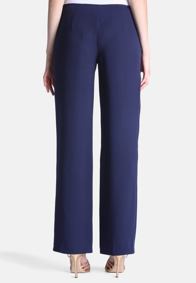 Personne portant un pantalon large bleu marine et des sandales à talons beiges, vue de dos sur un fond uni.
