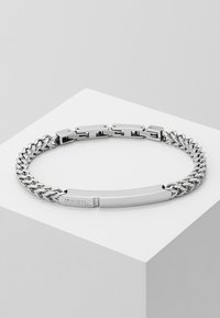 Stříbrný nerezový ocelový náramek s pleteným řetízkovým designem, plochou částí a detailem loga. Hladký povrch s metalickým leskem.