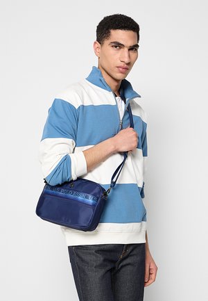 Borsa a tracolla blu navy in nylon con texture liscia, caratterizzata da una fascia con logo blu e tracolla regolabile. Design rettangolare compatto.