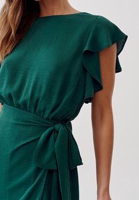 Abito verde con maniche a volant, vita cintrata con una fascia annodata e una texture di tessuto liscia e fluida.