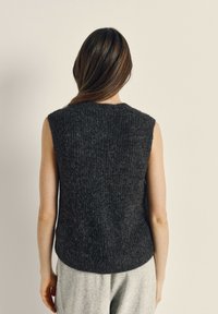 Mouwloos donkergrijs gebreid vest met een gestructureerde afwerking, afgeronde zoom en een losse pasvorm, gedragen over lichtgrijze broeken.