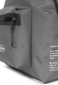 Šedá taška Eastpak má strukturovaný povrch, přední zipovou kapsu a bílé poti štěné texty, které zdůrazňují hodnoty značky a udržitelnost.
