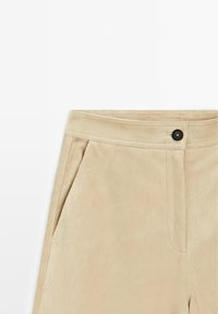 Beige tailor-made broek van zacht suede-achtig materiaal met een platte voorkant, twee zijzakken en een enkele zwarte knoopsluiting.