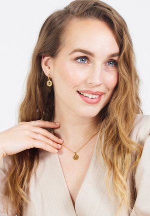 Goldene Ohrringe und eine passende Halskette mit runden Anhängern, die den Buchstaben 'M' zeigen. Der Schmuck wird mit einem leichten, strukturierten Oberteil getragen.