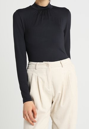 Kobieta ubrana w dopasowaną czarną bluzkę z długim rękawem i wysokim kołnierzem oraz kremowe, plisowane sztruksowe spodnie z wysokim stanem.