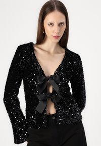 Crna jakna s sequinom, dugih rukava, V-izrezom i dva velika crna satenska mašna s prednje strane, s prilagođenim siluetom.