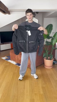 Young man standing indoors holding a black zip-up jacket with large front pockets and a white tag, next to a large potted plant.