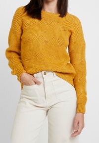 Hořčicově žlutý pletený svetr s vlnkovým vzorem, kulatým výstřihem a žebrovanými manžetami, v kombinaci se světlými kalhotami s vysokým pasem.