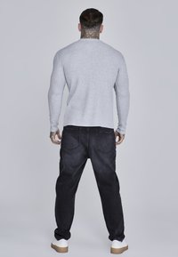 Szürke hosszú ujjú póló, kerek nyakkivágással, sötét farmernadrággal párosítva. A lábbeli világos színű, kontrasztos talppal rendelkezik.