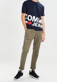T-shirt di cotone blu navy con grafica bianca "TOMMY JEANS". Pantaloni verde oliva con taglio dritto. Sneakers nere con accenti bianchi e rossi.