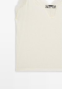 Haut sans manches de couleur crème avec un passepoil en dentelle au niveau du col. Tissu doux avec une texture lisse, comportant une étiquette au niveau du col.