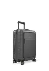Valise rigide grise avec des rainures verticales, poignée rétractable, poche zippée à l'avant et quatre roues pour la mobilité. Design durable.