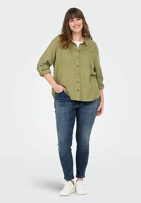 Camisa verde oliva de botones con bolsillos en el pecho, mangas enrolladas, acompañada de jeans ajustados azul oscuro y zapatillas blancas.
