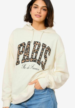 Krem hoodie s natpisom "PARIS" u uzorku leoparda, s kapuljačom na vezanje i kengur džepom. Uparen s plavim trapericama.