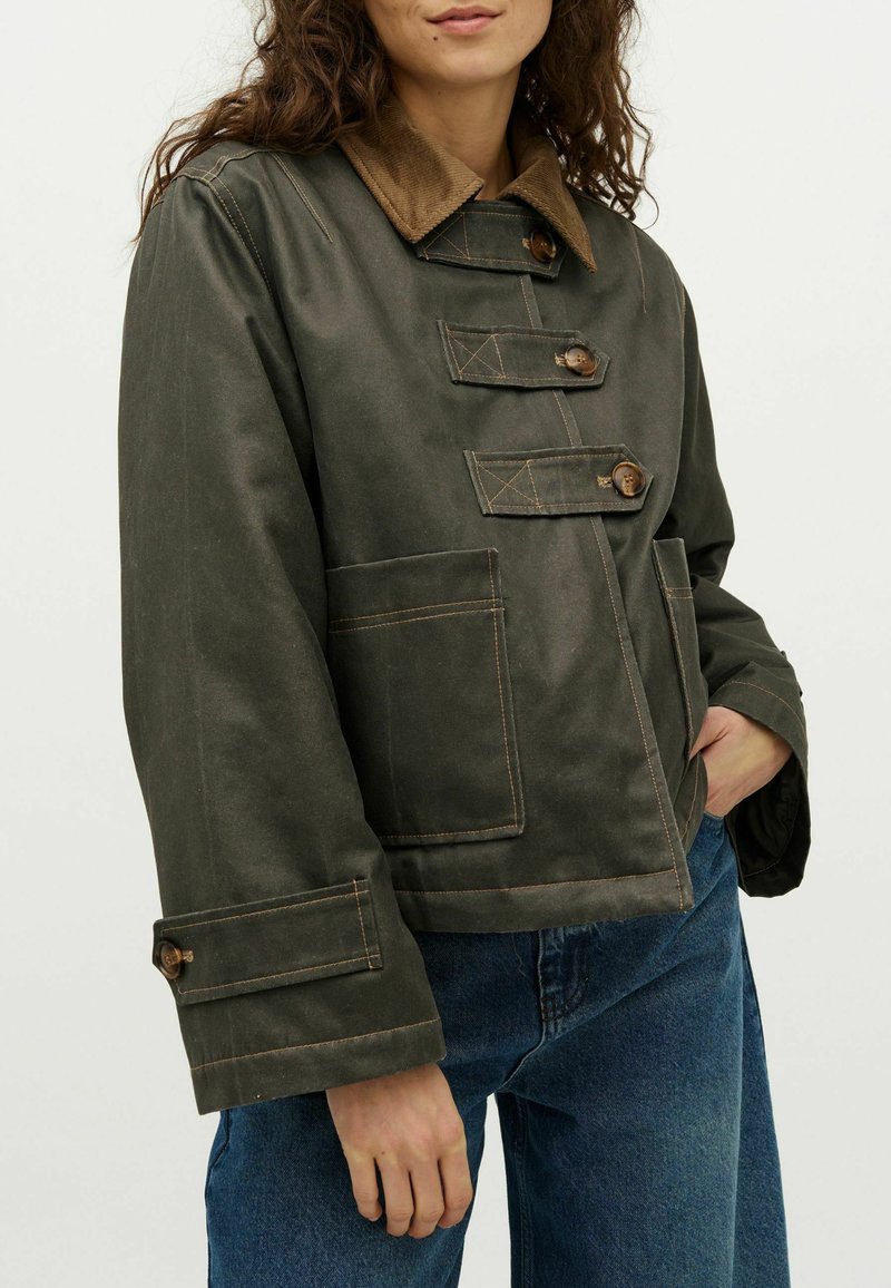 Nő sötétzöld dzsekiben, amelyen három gombos pánt található, foltvarrott zsebekkel és barna kordbársonnyal, kék farmernadrággal párosítva.