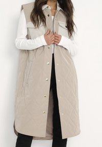 Beige gesteppte Weste mit Druckknöpfen, ärmellosem Design, Rautenmuster und zwei vorderen Taschen, getragen über einem weißen Langarmtop.