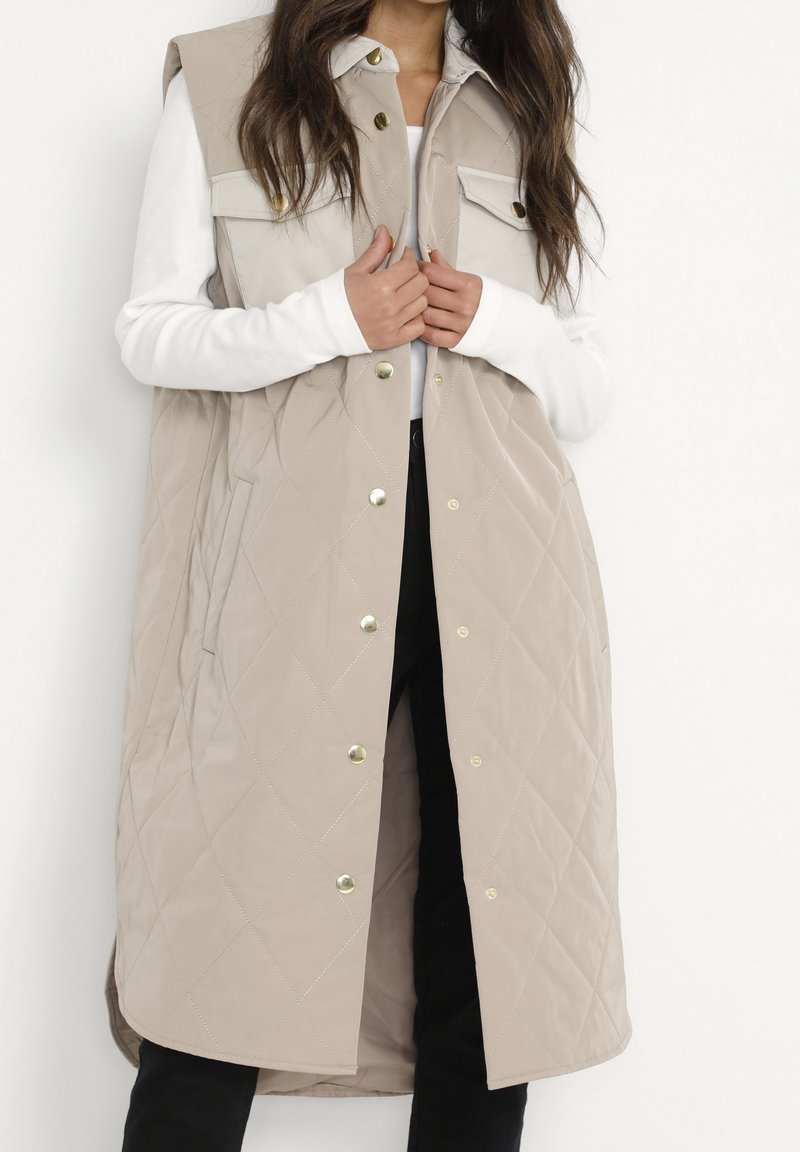 Beige gesteppte Weste mit Druckknöpfen, ärmellosem Design, Rautenmuster und zwei vorderen Taschen, getragen über einem weißen Langarmtop.