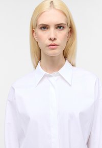Hvid skjorte med krave og lange ærmer, lavet af glat stof, med en knap-lukning foran og en afslappet pasform. Rent, minimalistisk design.